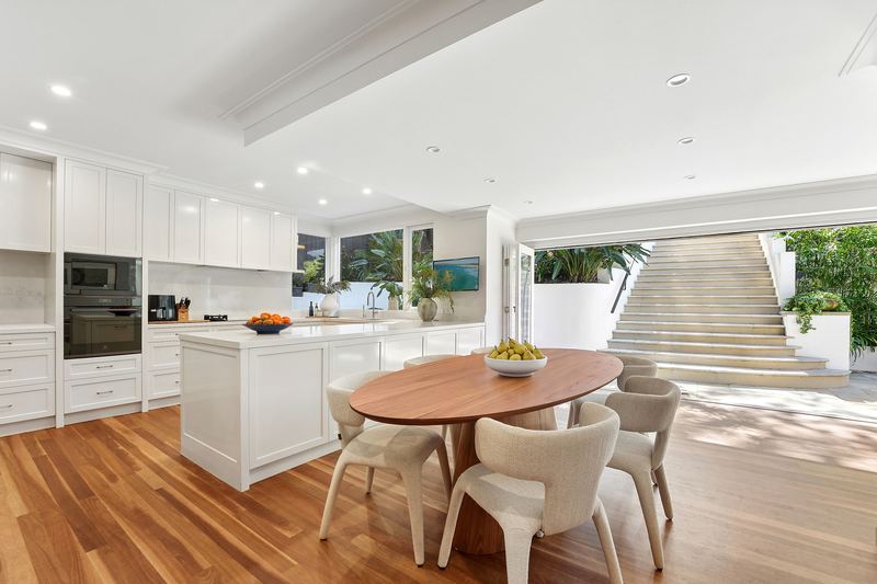 White kitchen with island and oval timber dining table, doors open to the terrace and garden stairs