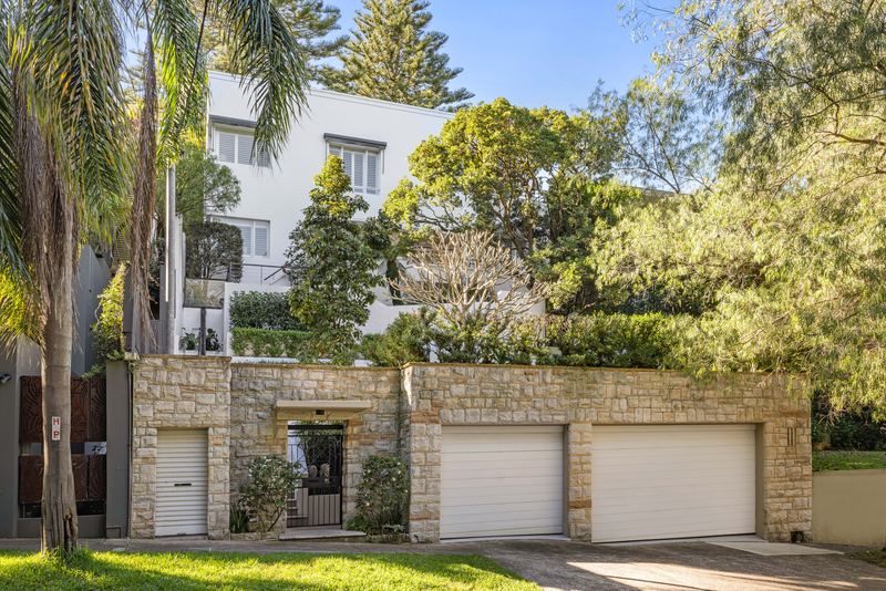 Street frontage with a sandstone wall, three garage doors and the white rendered house above among palms and trees
